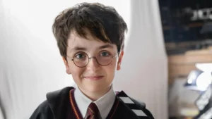A young boy dressed as Harry Potter, wearing round glasses and a Hogwarts uniform, holding a clapperboard and smiling at the camera.